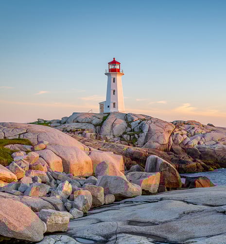 A white lighthouse with a red top stands atop rugged, sunlit rocks by the sea under a clear blue sky, evoking a serene and coastal ambiance.