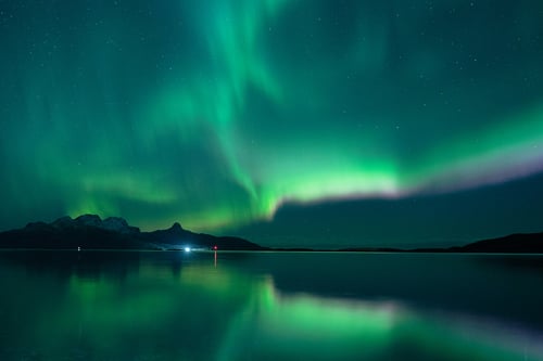 Vibrant green aurora borealis reflecting in calm water with distant mountains and lights.