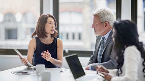 A diverse group of three professionals in a meeting room, with laptops and papers on the table. The woman in black speaks passionately.