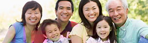 A joyful multi-generational family of six, including grandparents, parents, and two children, smiles at the camera outdoors on a sunny day.