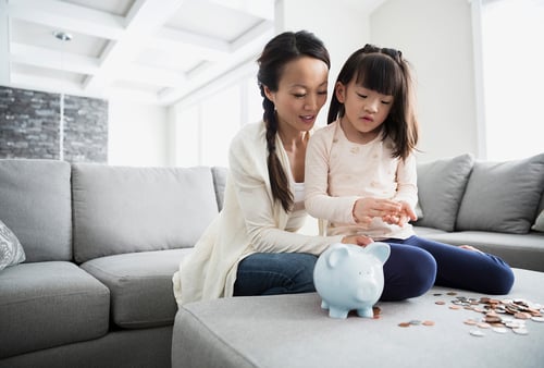 A woman and young girl sit on a gray sofa, counting coins beside a blue piggy bank. The room is bright and modern, conveying a warm, educational moment.
