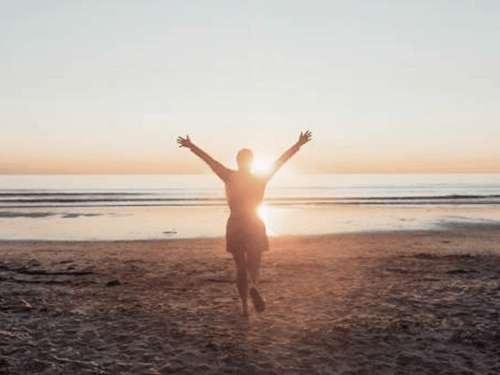 A woman running with her arms raised on a beach, at sunset.