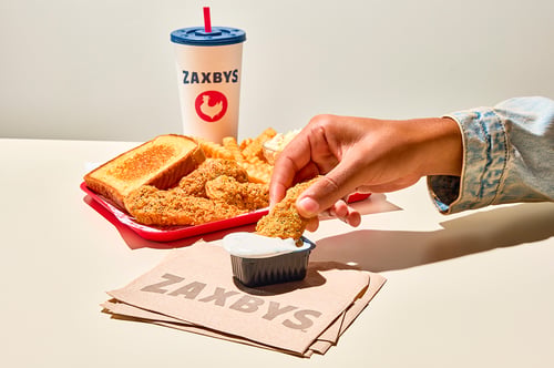Hand dipping a Zaxby's Chicken Finger into Ranch sauce with Texas toast, crinkle fries, coleslaw, and a drink in the background.