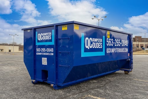 Blue QC Dumpster Dudes dumpster parked in an empty lot, featuring company branding and contact details.