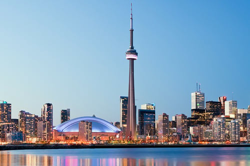 Toronto skyline at dusk with the CN Tower prominently lit. Skyscrapers surround it, reflecting vibrant lights on calm water, creating a serene urban scene.
