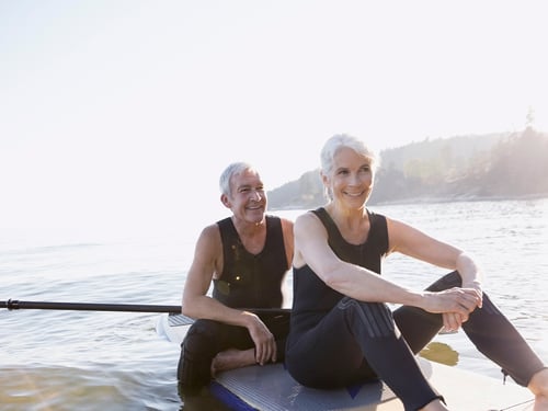 A smiling older couple in wetsuits sit on paddleboards on a calm lake, bathed in sunlight. The scene conveys tranquility and joy.