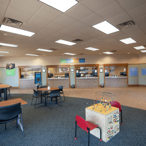 Four teller stations line the back wall in the photo. A sitting area with several tables and chairs are set up across the room from the counters. There is also a small play area for children next to the other tables.