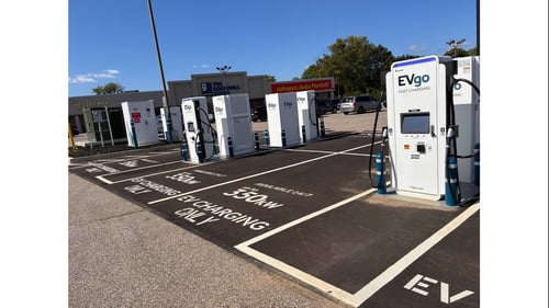 Several EVgo fast charging units installed in marked parking spaces at a public charging station, with EVgo logos on the chargers, protective bollards around each unit. CCS chargers featured in the image.