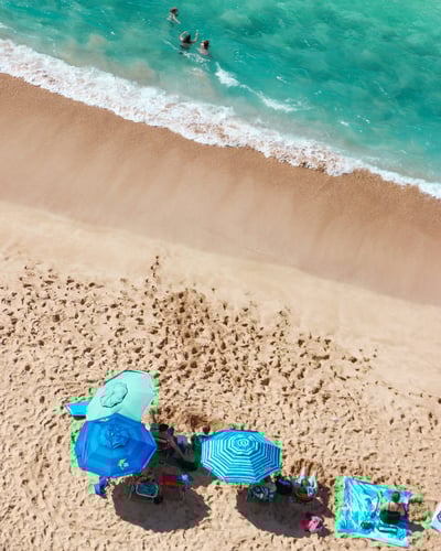Aerial view of pristine beach with three swimmers in crystal-clear turquoise water, beachgoers relaxing under blue striped umbrellas on golden sand with gentle white waves at shoreline.