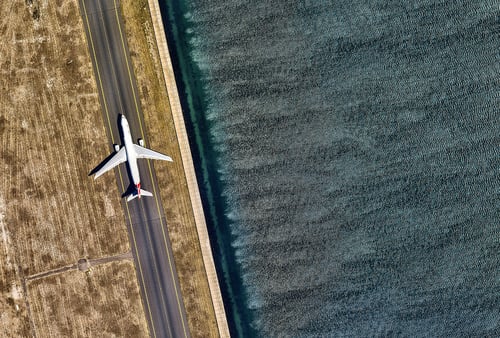 Aerial view of an airplane taxiing on a narrow runway beside shimmering water on the right. Dry, brown grass covers the left side, contrasting the smooth surface.