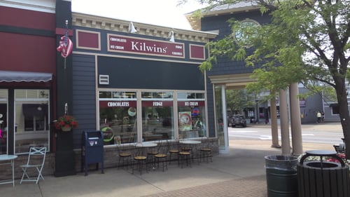 Exterior of the Kilwins Boyne City, MI 49712 chocolate, fudge, and ice cream store with round tables and chairs for seating outside of the storefront