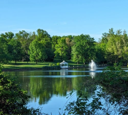A lake on the grounds with a fountain playing and a white gazebo on the far bank, framed by mature trees