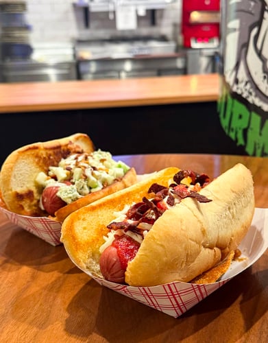Two gourmet hot dogs with homemade potato chips in red plaid to-go tray. Dead Dixie Dog (left) and Blood Graffiti Dog (right).