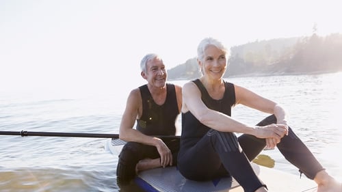 A smiling older couple in wetsuits sit on paddleboards on a calm lake, bathed in sunlight. The scene conveys tranquility and joy.