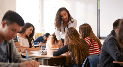 A teacher assists a small group of engaged students in a brightly lit classroom. Other students are focused on their work, creating a collaborative atmosphere.