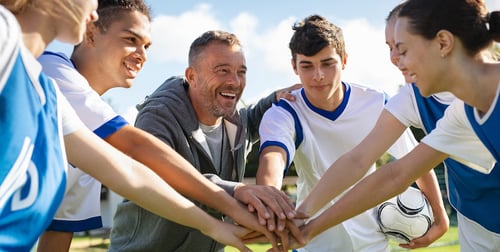 "Happy coach and high school soccer team with their hands stacked in football court. Young football players stacking hands together. Mature man coach encourages his students to do their best during the sport match. (Happy coach and high school soccer t"
