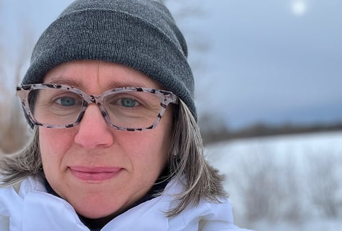 Colleen wearing glasses, a gray beanie, and a white jacket stands outdoors in a snowy landscape. The overcast sky sets a calm, chilly mood.