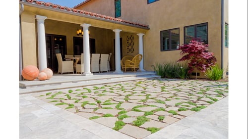 Sunlit, inviting Mediterranean courtyard with covered patio, white columns, outdoor dining, wicker chair, patterned stone paving, and greenery.