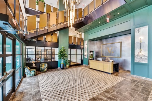 A lobby with green walls, a gold reception desk, glass doors, a brass and wood staircase wrapping around the perimeter, and a modern chandelier.