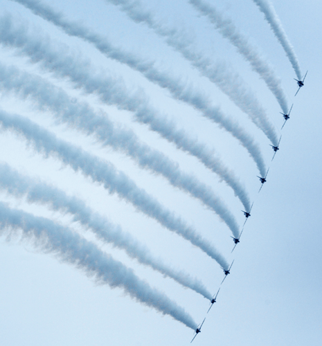 Nine jet planes fly in formation against a clear blue sky, leaving thick, curved trails of white smoke. The scene conveys precision and unity.