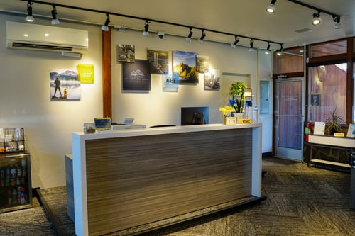 The interior of a bank lobby featuring a modern reception desk with a computer monitor. The wall behind the desk is decorated with framed pictures, including one with the "Idaho First Bank" logo. Overhead track lighting illuminates the space.