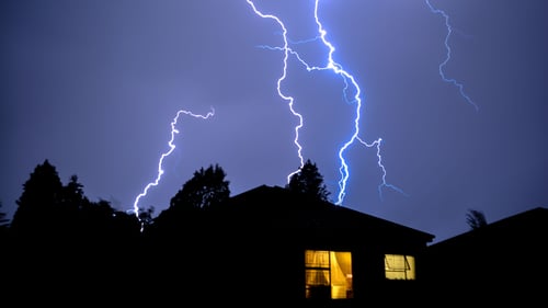 Lightning lights up the sky behind a home with lights coming through the windows.