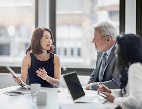 A diverse group of three professionals in a meeting room, with laptops and papers on the table. The woman in black speaks passionately.