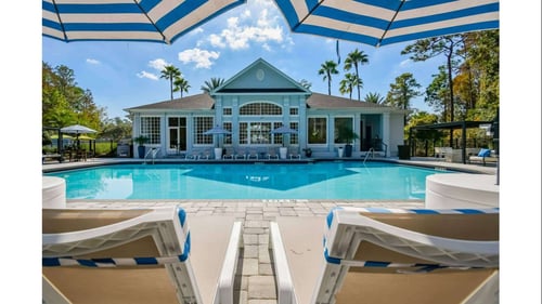 A ground-level view across the swimming pool toward the clubhouse building, framed by lounge chairs and blue-and-white striped umbrellas in the foreground.