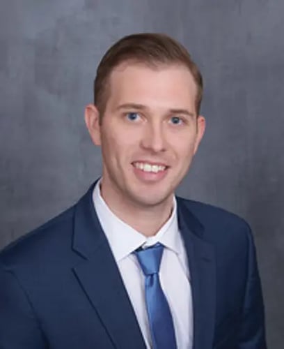 Agent Ryan Bush sitting and smiling wearing white button up shirt with blue tie and navy blue blazer