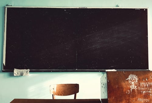 An empty classroom with a large black chalkboard on a teal wall. A solitary wooden chair and a desk with doodles add a nostalgic, quiet atmosphere.