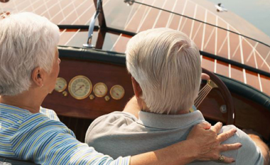 Elderly couple with white hair on a boat, seen from behind. Woman rests her arm on man's shoulder. Wooden deck and water in background, conveying tranquility.