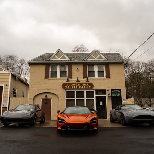 Exterior photo of Flower Hill Auto Body in Roslyn, New York, showing the front of the building with the Flower Hill Auto Body sign and three luxury vehicles parked in front, including a sports car and high end sedans, on a wet overcast day.