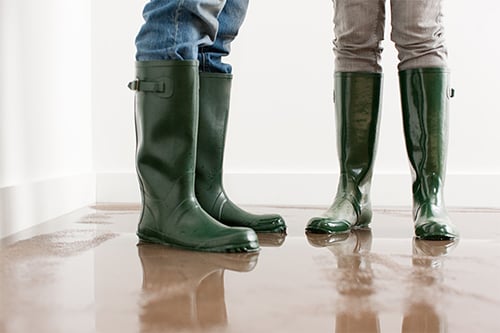 Two homeowners stand in a water damage puddle while wearing green boots.
