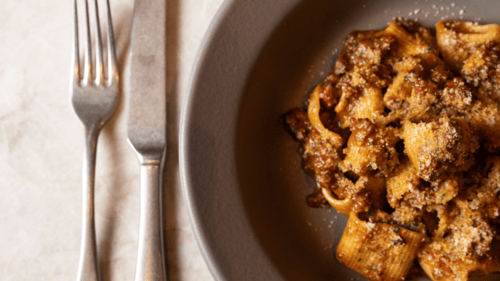 Plate of rigatoni pasta with meat sauce and grated cheese next to a fork and knife on a marble surface