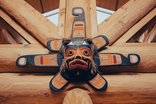 A large traditional Pacific Northwest Coast Indigenous carved wooden sun mask mounted against a ceiling framework of massive natural logs, featuring a 3D painted face surrounded by bark fringe and radiating wooden panels with black and orange-red formline designs.