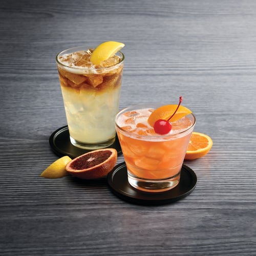 Two cocktails on a wooden surface, one tall layered iced drink with lemon garnish and one short orange-toned cocktail with cherry and citrus slices.