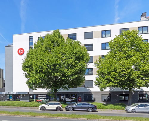 Modernes Gebäude mit Studentennapartments, weiße Fassade mit dunklem Erdgeschoss und großen Fenstern, davor mehrere geparkte Autos und dicht begrünte Bäume entlang einer mehrspurigen Straße unter blauem Himmel.