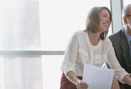 A woman in a white blouse and red skirt smiles brightly while holding papers, next to a blurred person in a suit, against a bright window backdrop.