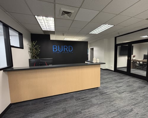Office reception with BURD Home Health sign, wood desk, gray carpet, and glass-walled rooms.