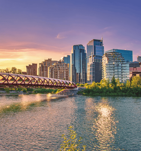 A vibrant cityscape at sunset features a river and a distinctive red, lattice bridge in the foreground, set against modern skyscrapers and a colorful sky.