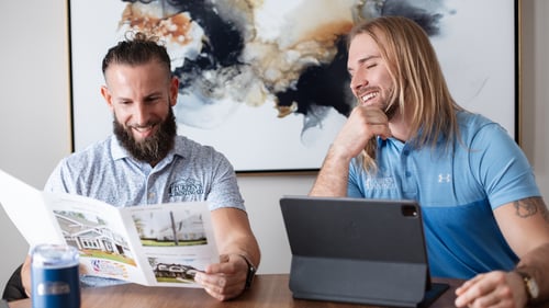 Two men smiling and sitting at a table, one reading a brochure, the other looking at a tablet. Abstract art in the background creates an engaging atmosphere.