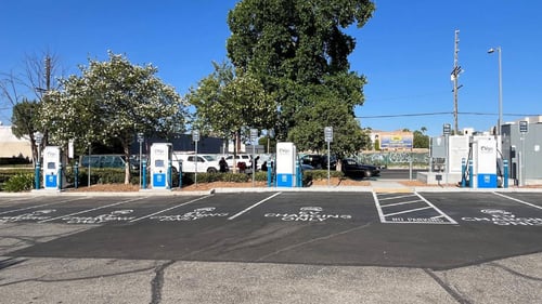Several EVgo fast charging units installed in marked parking spaces at a public charging station, with EVgo logos on the chargers, protective bollards around each unit. Front view.