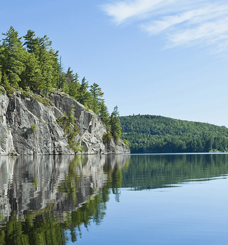 A calm lake with a rocky cliff and a lush forested mountain in the background.