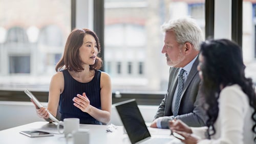 A diverse group of three professionals in a meeting room, with laptops and papers on the table. The woman in black speaks passionately.