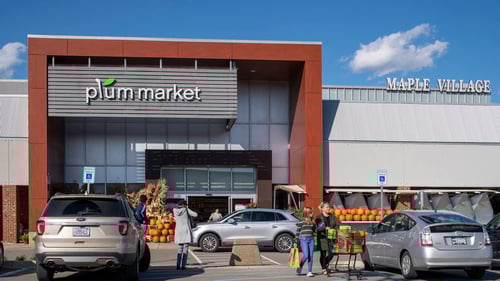 People with shopping carts walking in front of pumpkin lined entrance of Plum Market