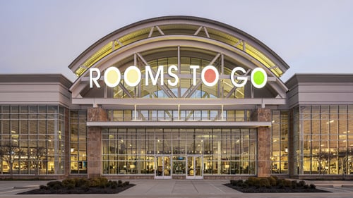 Rooms To Go store facade at dusk in The Colony, TX, featuring large glass windows and prominent illuminated signage.