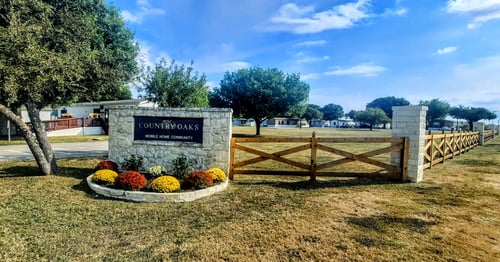 A stone sign that says "Country Oaks Mobile Home Community" stands in a grassy area at Country Oaks Logo in San Antonio, TX
