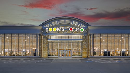 Front view of Rooms To Go store in Rockwall, TX, at twilight, with the store's name brightly lit and large parking area.