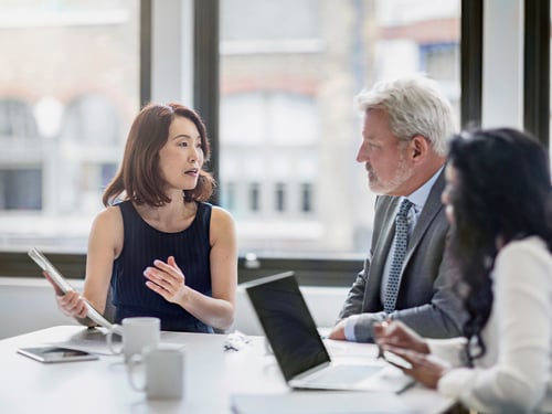 A diverse group of three professionals in a meeting room, with laptops and papers on the table. The woman in black speaks passionately.