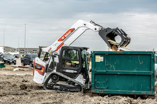 Operator is dropping placing wood into a dumpster with a Bobcat T64 equipped with an industrial grapple